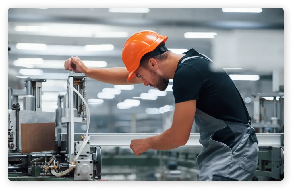 A worker is wearing an orange hard hat, black t-shirt and work overalls whilst using a heavy duty machine.