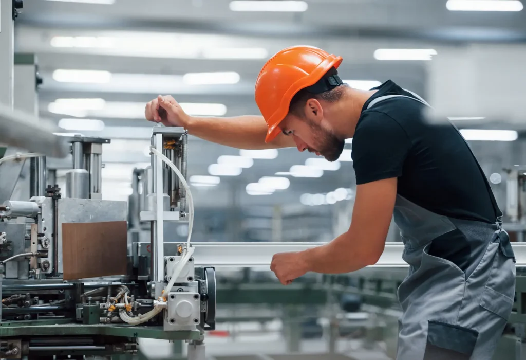 An individual wearing a orange hard hat, a black t-shirt and work overalls is using a heavy duty machine.
