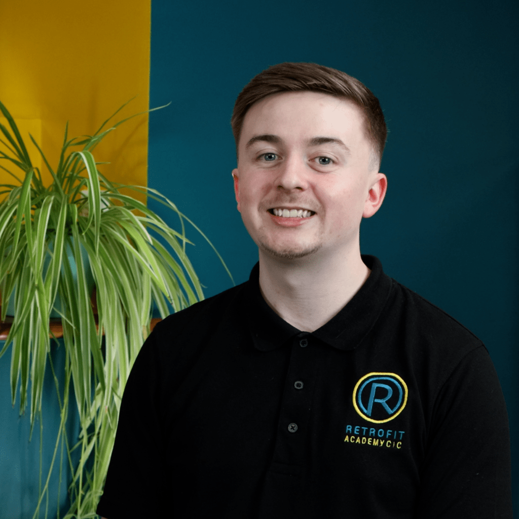 An individual called Charlie Hayes is wearing a Retrofit branded polo whilst posing next to a blue and yellow wall and a plant.