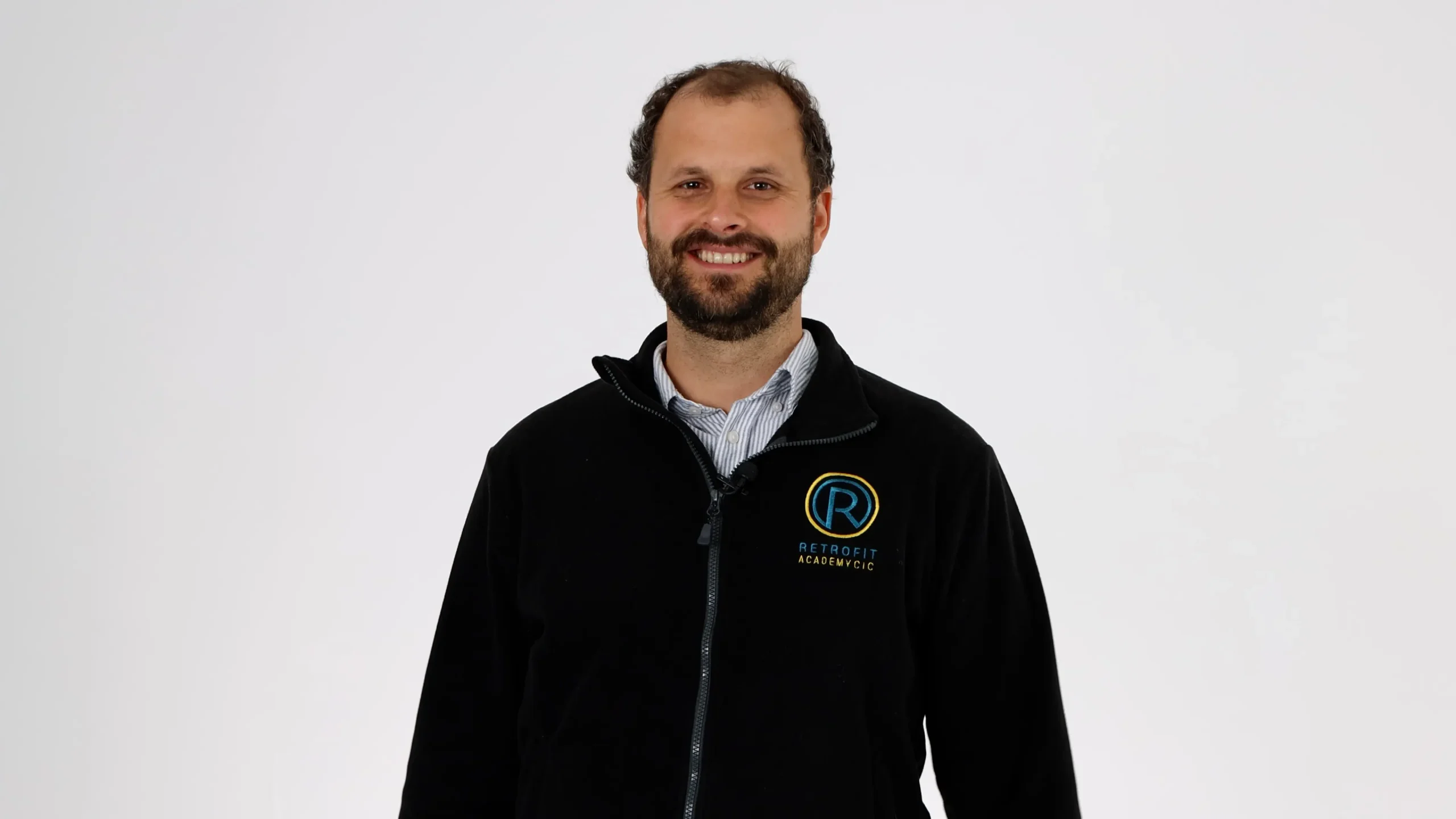 A photo of an individual called Dom Ives who has short brown hair and a beard is wearing a black Retrofit Academy jacket whilst smiling at the camera.