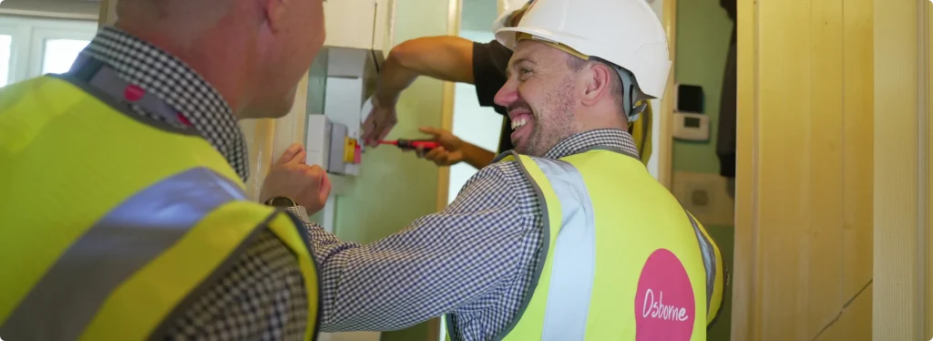 Two individuals wearing white hard hats, checked shirts and yellow hi-vis vests are working on fixing an electric box inside a house.