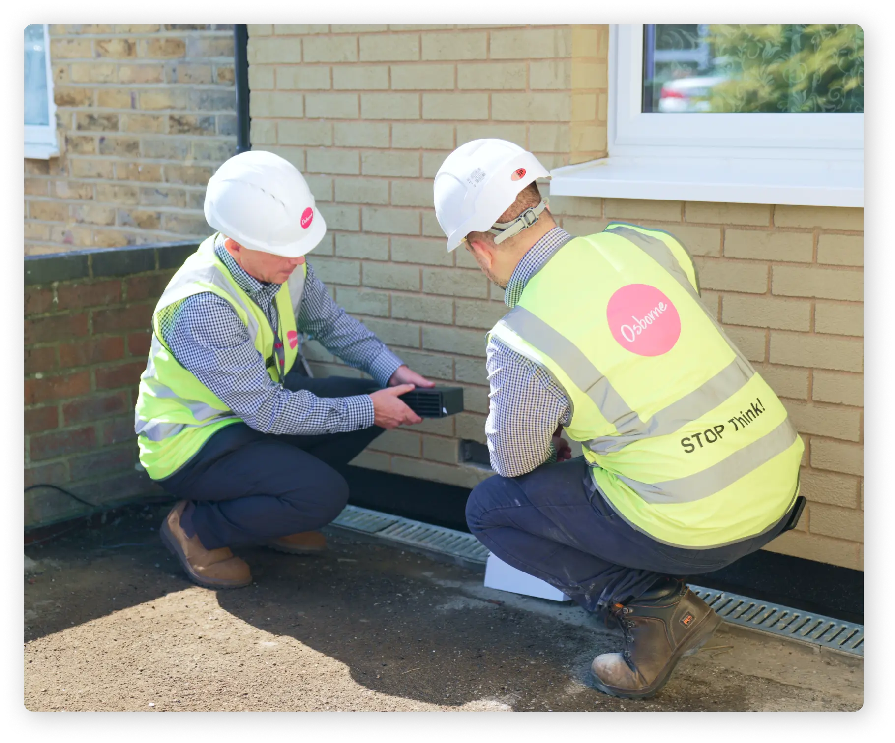 Two construction workers who are wearing white hard hats, checked shirts and hi-vis vests are installing vents into a brick house.