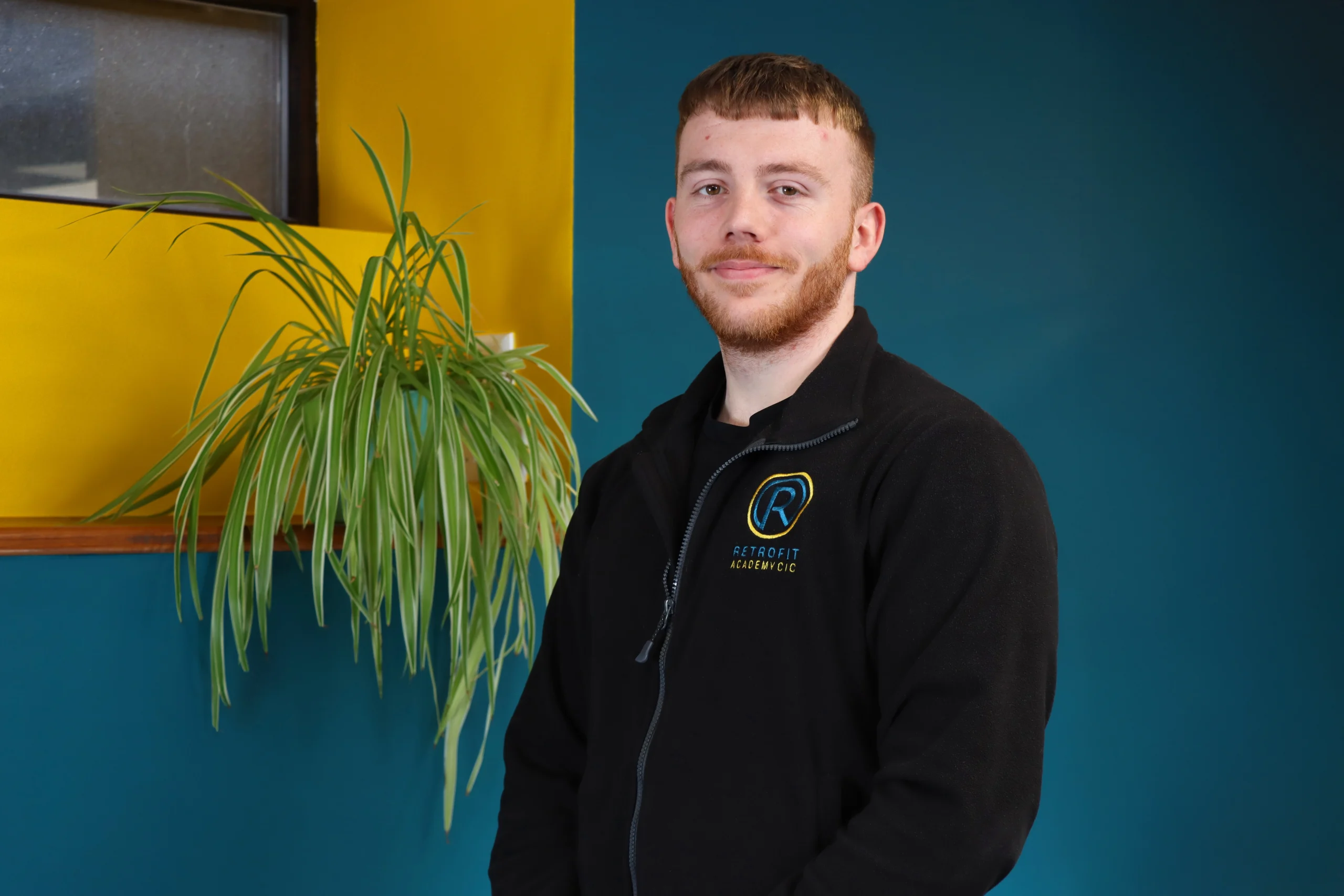 A photo of Josh, a member of the Retrofit Academy team, is wearing a branded black Retrofit Academy zip up jacket, has short brown hair with brown facial hair and is standing in front of blue and yellow walls with a green plant next to him.