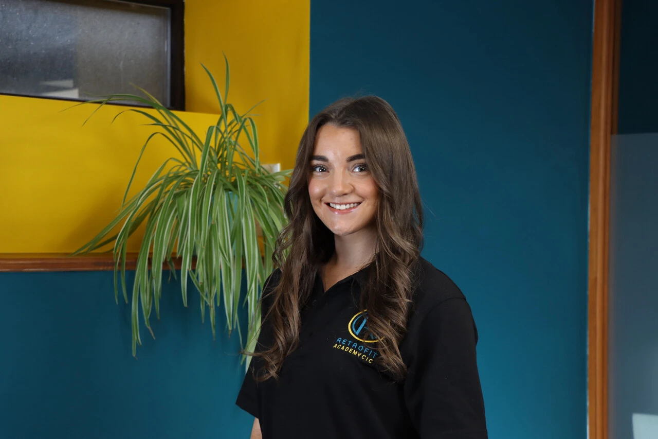 An individual called Katie Felton is posing in front of a yellow and blue wall next to a plant, whilst wearing a black Retrofit Academy branded polo shirt.