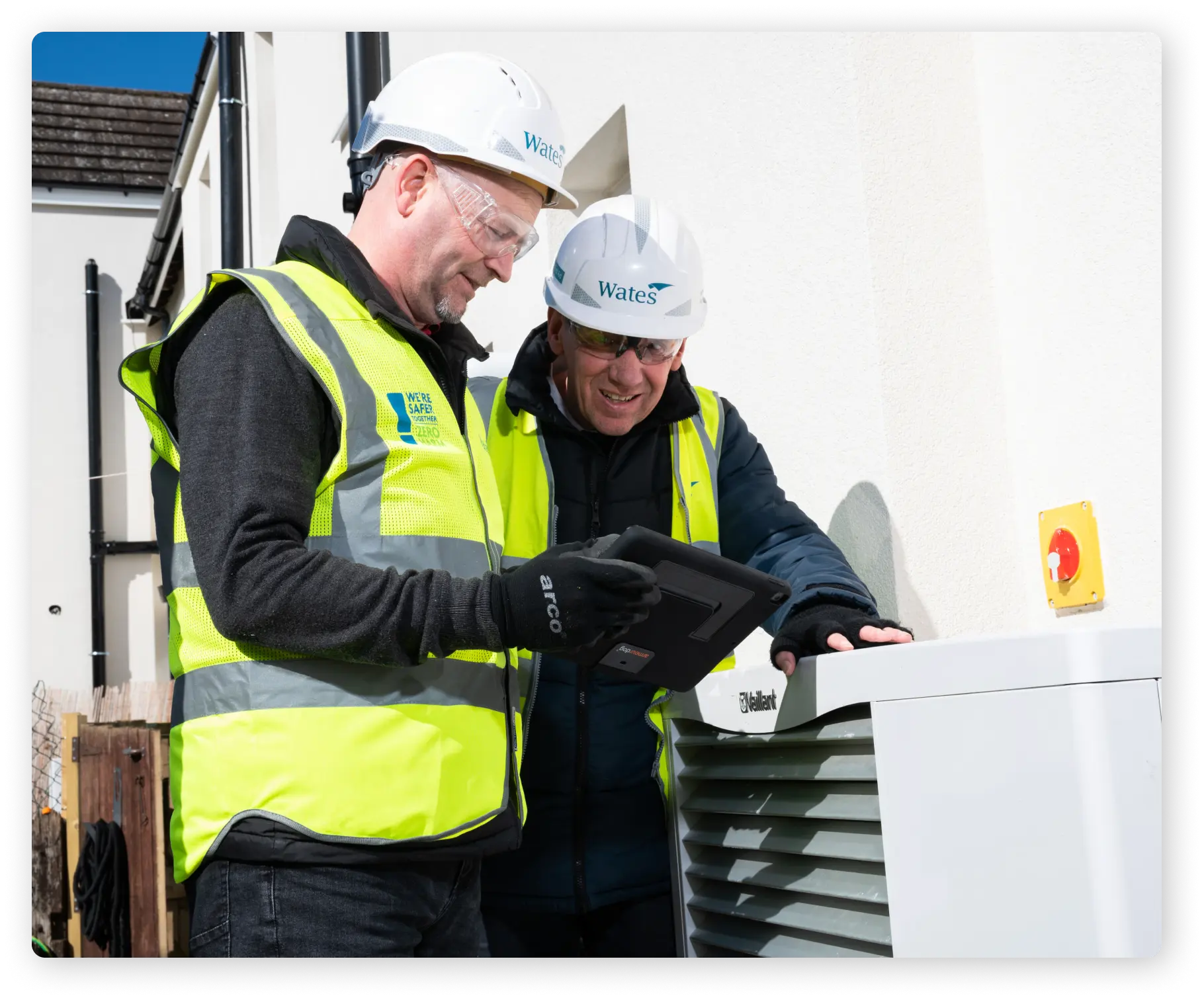 Two construction workers wearing white hard hats and hi-vis vests are looking at the screen of a black tablet.
