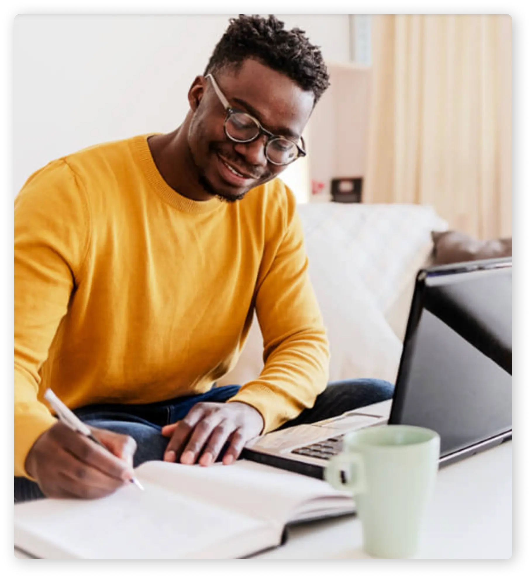 An individual wearing a yellow jumper, blue jeans and glasses is sat at a table writing in a notebook with a laptop in front of them.