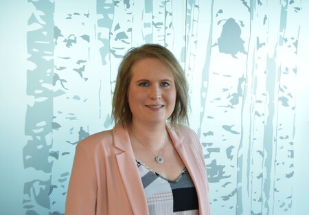 An individual who has short shoulder length brown hair is wearing a pink blazer with a black and white patterned top and is standing in front of a wall with a fake tree pattern.