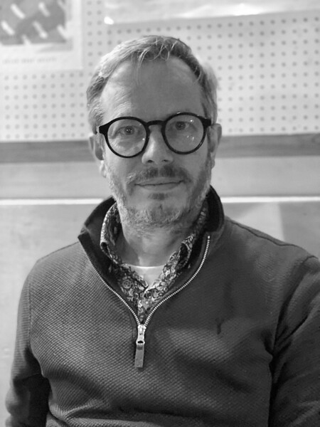An individual who has short grey hair and beard is wearing a jumper whilst smiling at the camera.