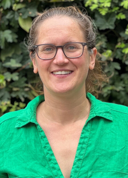 An individual called Lizzy Westmacott is standing in front of a green bush whilst wearing brown glasses and a bright green shirt.