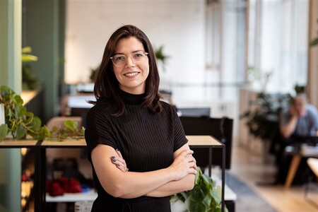 A photo of an individual who has long brown hair, clear glasses and a long black shirt, whilst posing in an office environment.