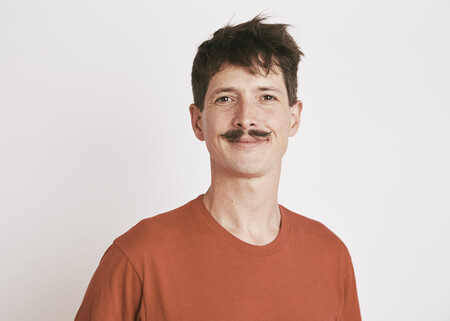 An individual who has short brown hair and a moustache whilst wearing a light orange t-shirt is smiling at the camera.