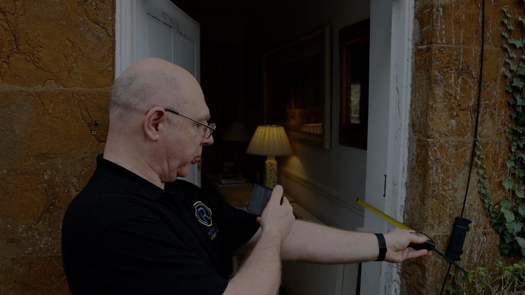An individual called Clive is using a yellow tape measure to measure the width of a door frame, whilst taking a picture of the measurement with their phone and wearing a black Retrofit Academy branded polo shirt.