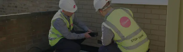 Two construction workers are wearing hard hats and hi-vis vests whilst working to fix a brick wall.