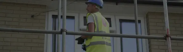 An individual wearing a blue hard hat, a yellow hi-vis vest and black gloves is standing in front of a house that has scaffolding up.
