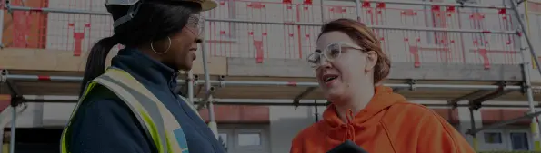 Two individuals one wearing a hard hat and a hi-vis vest the other wearing an orange hoodie are having a conversation in a construction site.