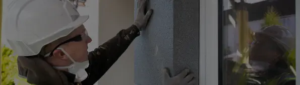 An individual wearing a white hard hat, a yellow hi-vis vest, a black hoodie and grey gloves is working on fixing a house.