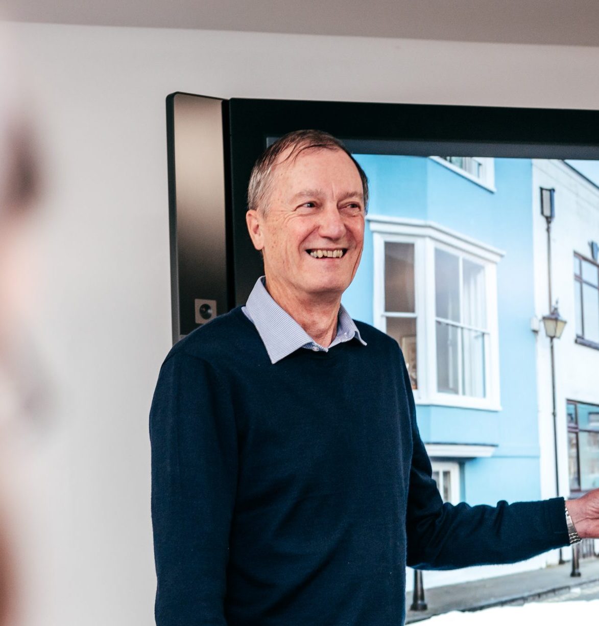 A photo of an individual who has short grey hair, is wearing a dark blue jumper and a light blue shirt, is smiling at a camera.