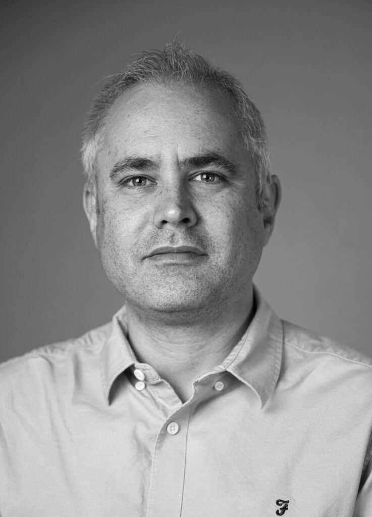 A black and white photo of Richard Fitton, who has short grey hair and is wearing a shirt.